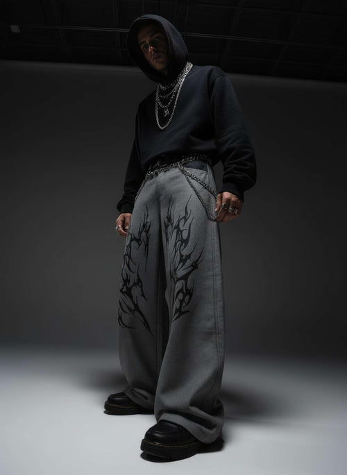 Person wearing a dark hoodie, light gray pants with black patterns, and black shoes in a minimalistic setting.