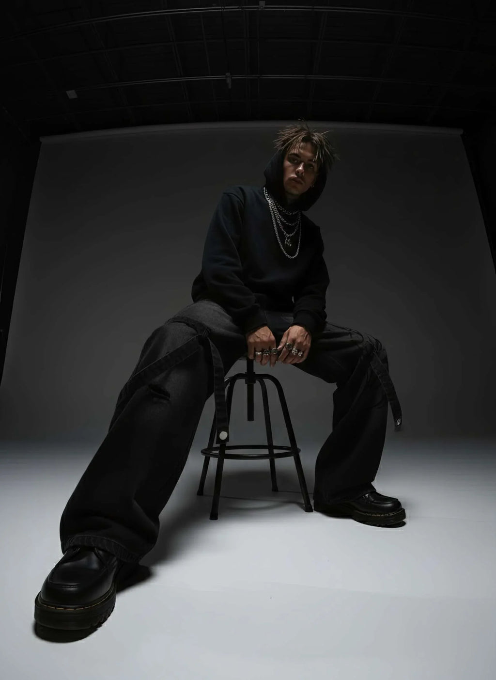 Person wearing a black outfit sitting on a stool against a dark background