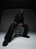 Person wearing a black outfit sitting on a stool against a dark background