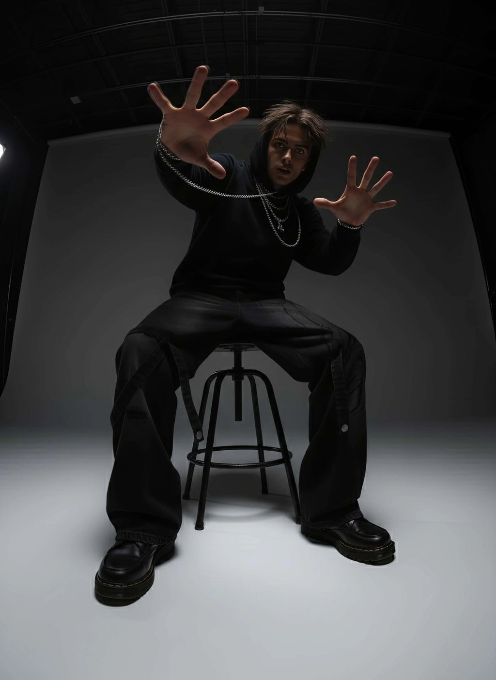 Person in all black sitting on a stool with arms outstretched in a dark studio setting